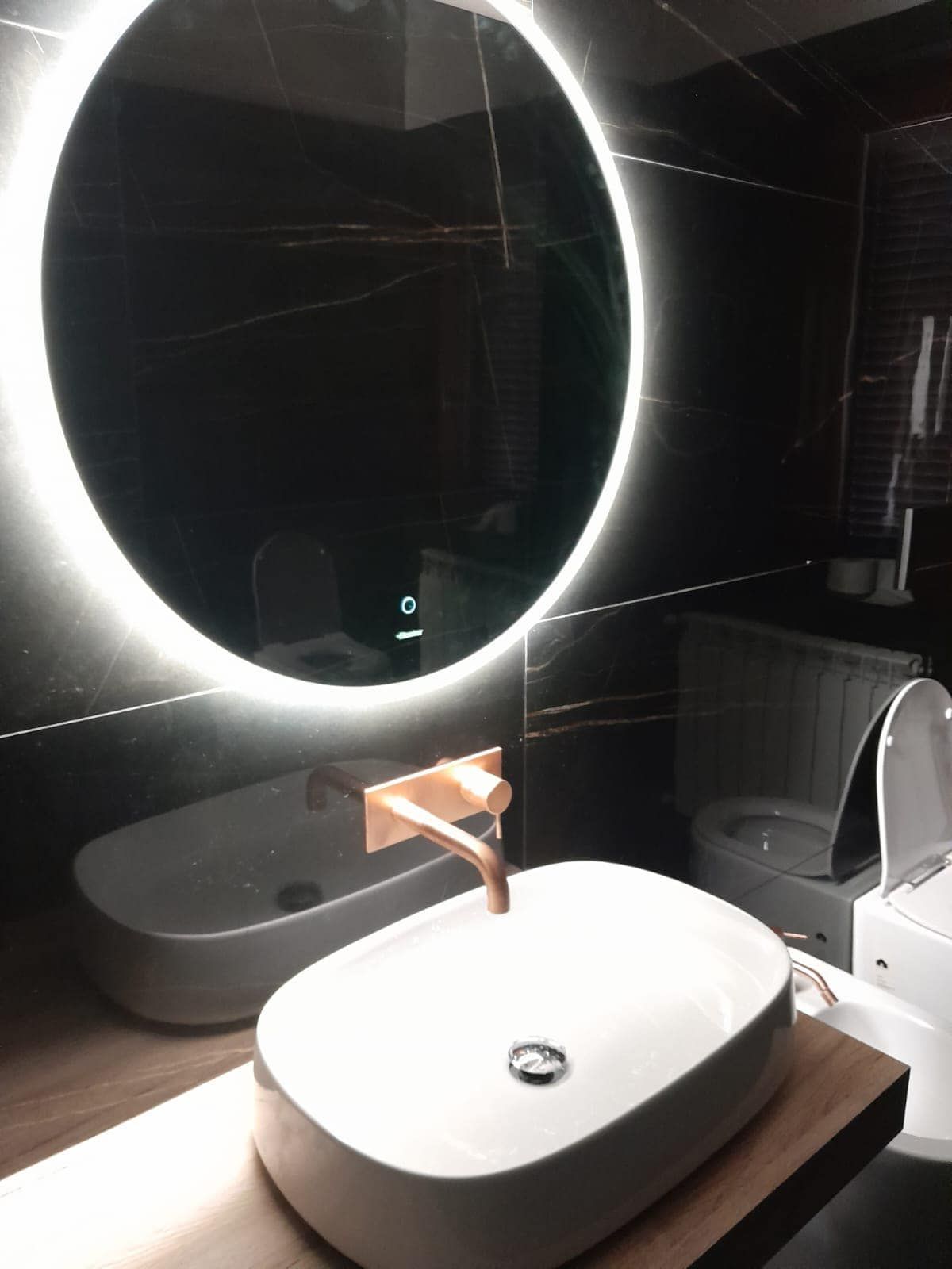 Bagno moderno con specchio circolare illuminato, lavandini bianchi e rubinetto in rame su piastrelle nere.