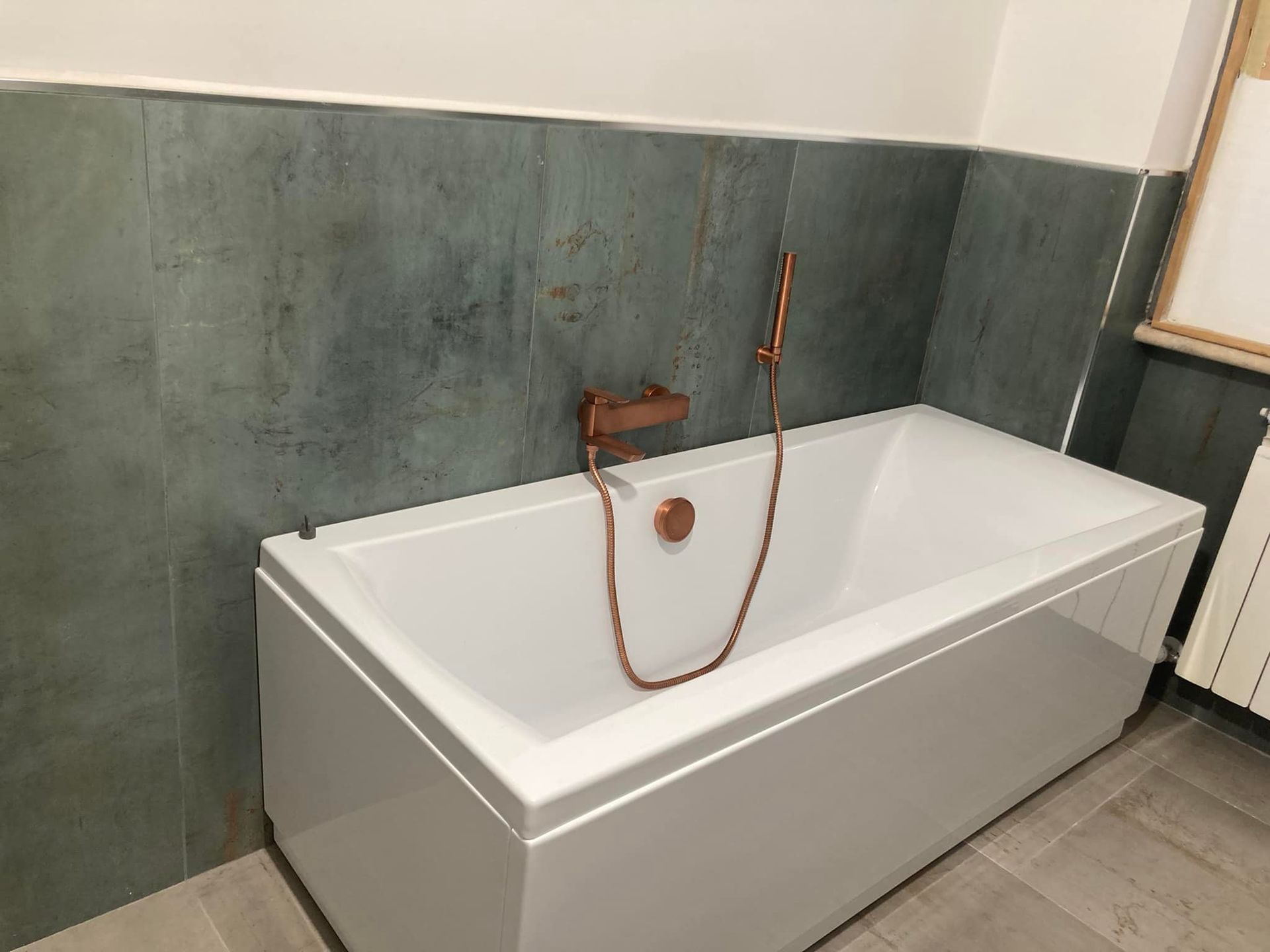 Vasca da bagno bianca con accessori in rame contro una parete piastrellata color verde acqua.
