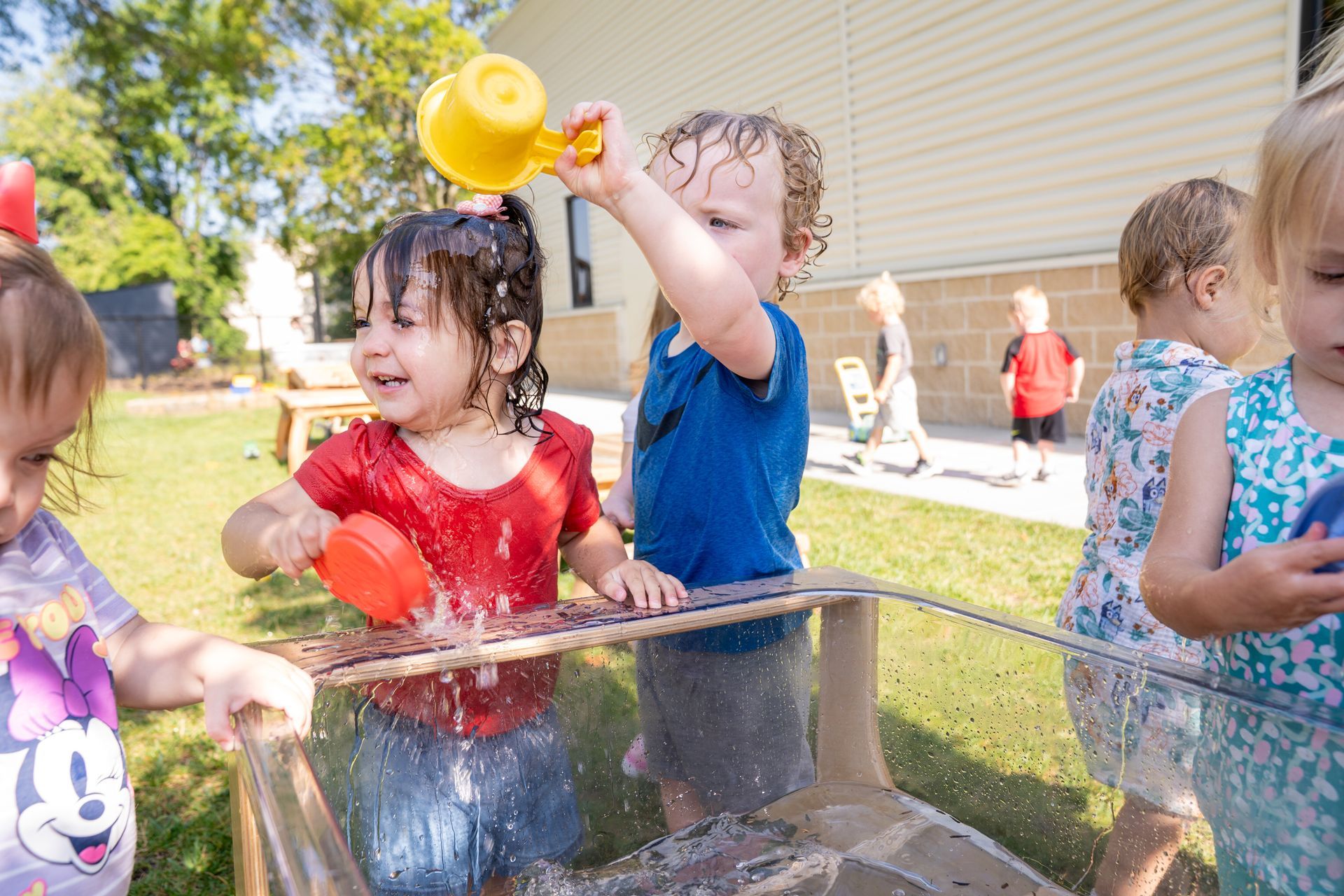 Children playing with water in a clear container outdoors. One is pouring water from a yellow cup, causing splashes.