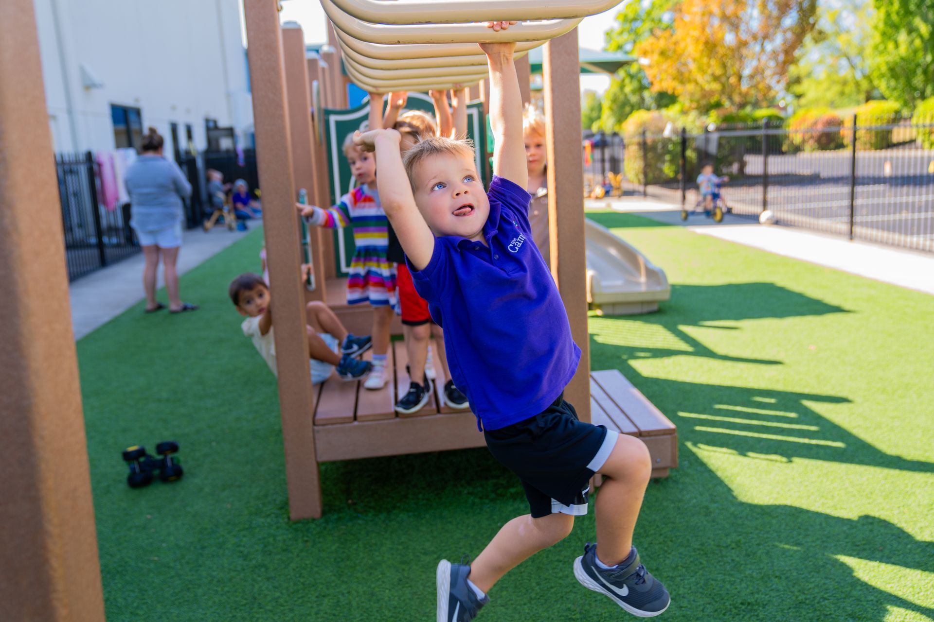 Boy in purple shirt hangs from monkey bars on a playground, other children play.
