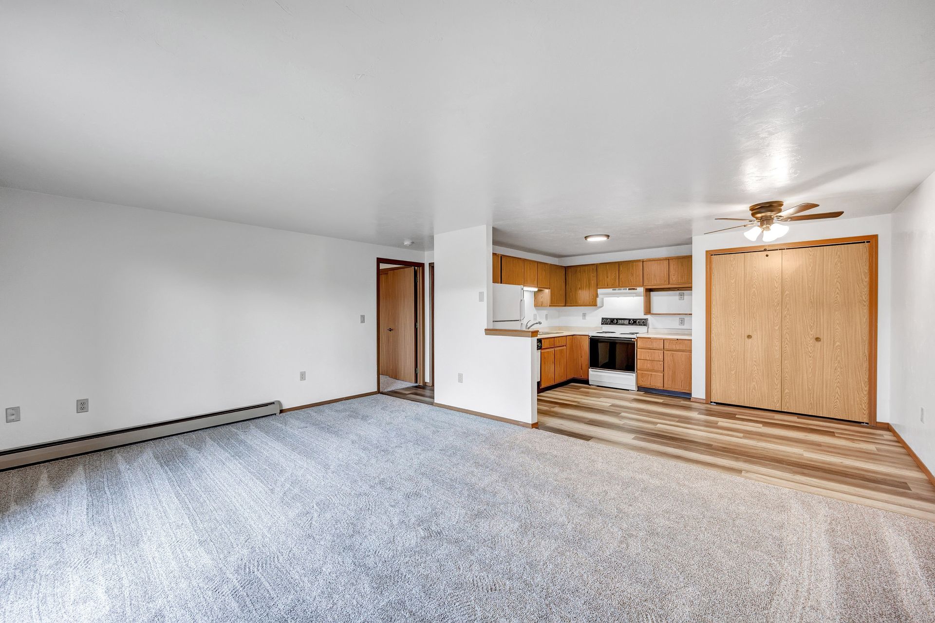 An empty living room with a ceiling fan and a kitchen in the background.