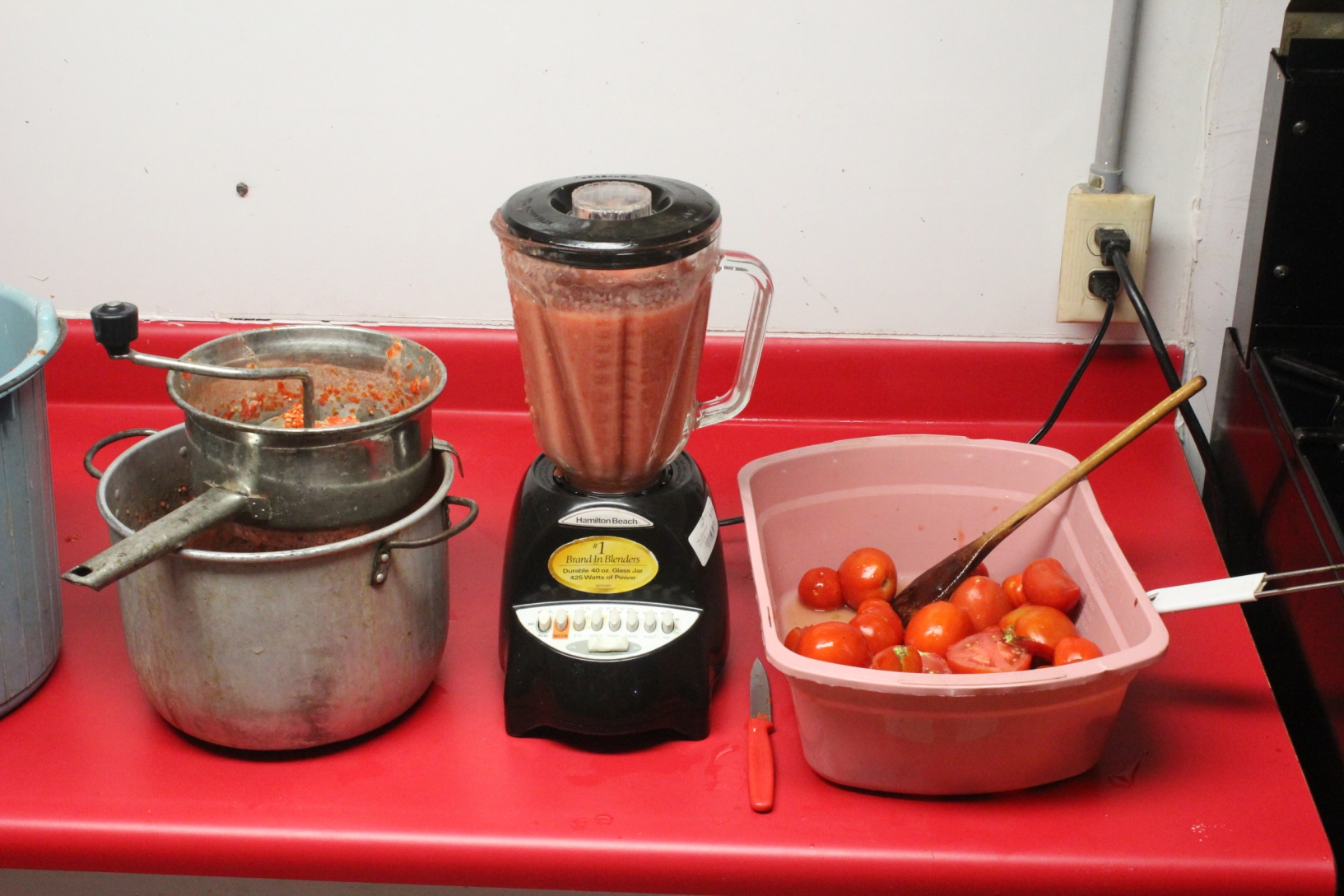 Photo of a container under the food mill which receives the tomato juice, a blender that pulverizes whole tomatoes and a plastic container holding cleaned tomatoes ready for processing