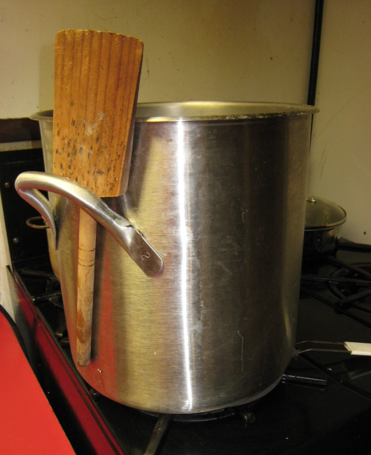 Photo of a large stainless steel pan on the stove with a wooden ladle. The tomato juice is cooking down into thick sauce.