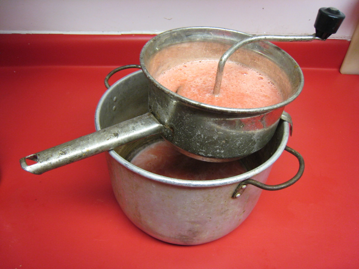 Photo of a food mill filled with pulverized tomatoes waiting to be spun and separated with the juice going into the pan below