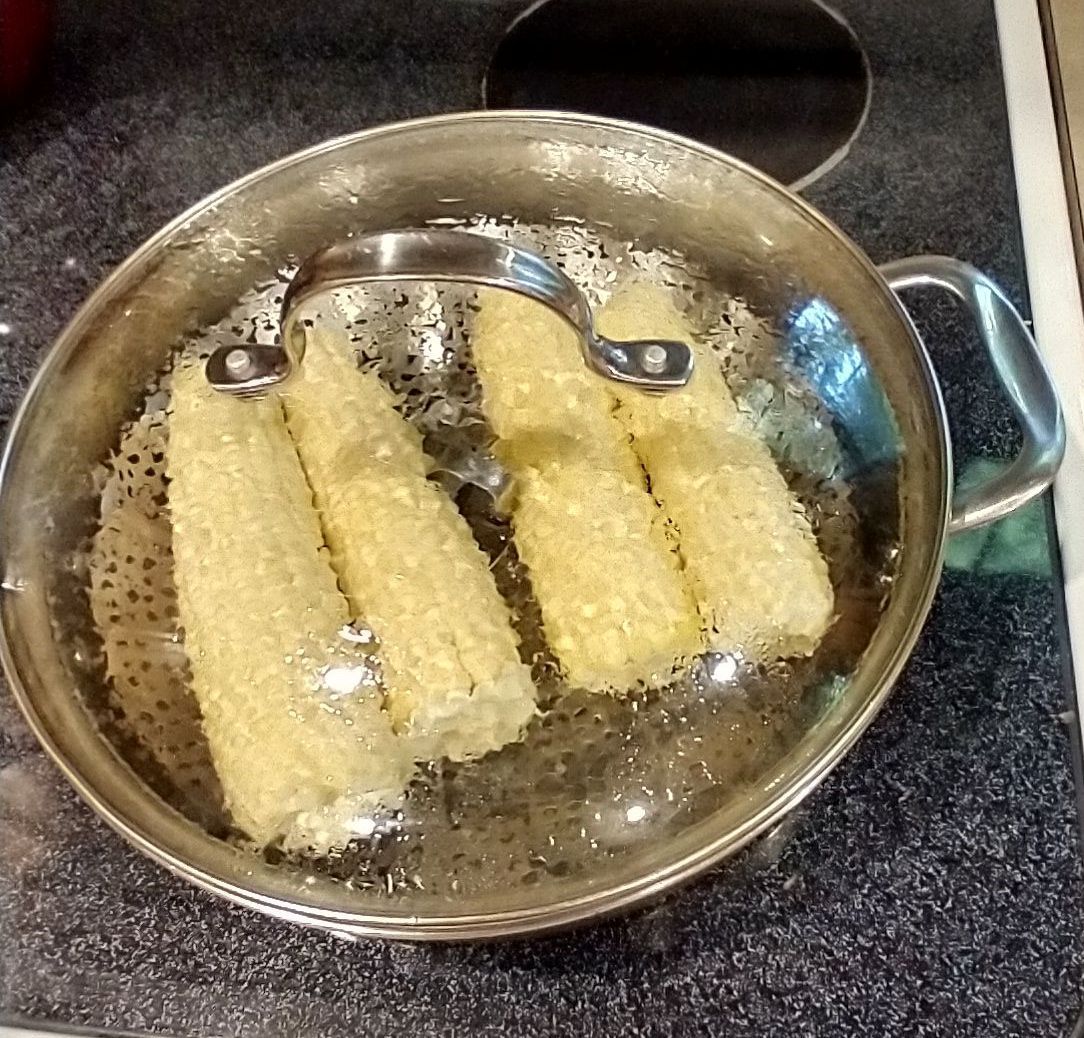 Four cobs of sweet corn in a steamer basket above simmering water in a closed frying pan