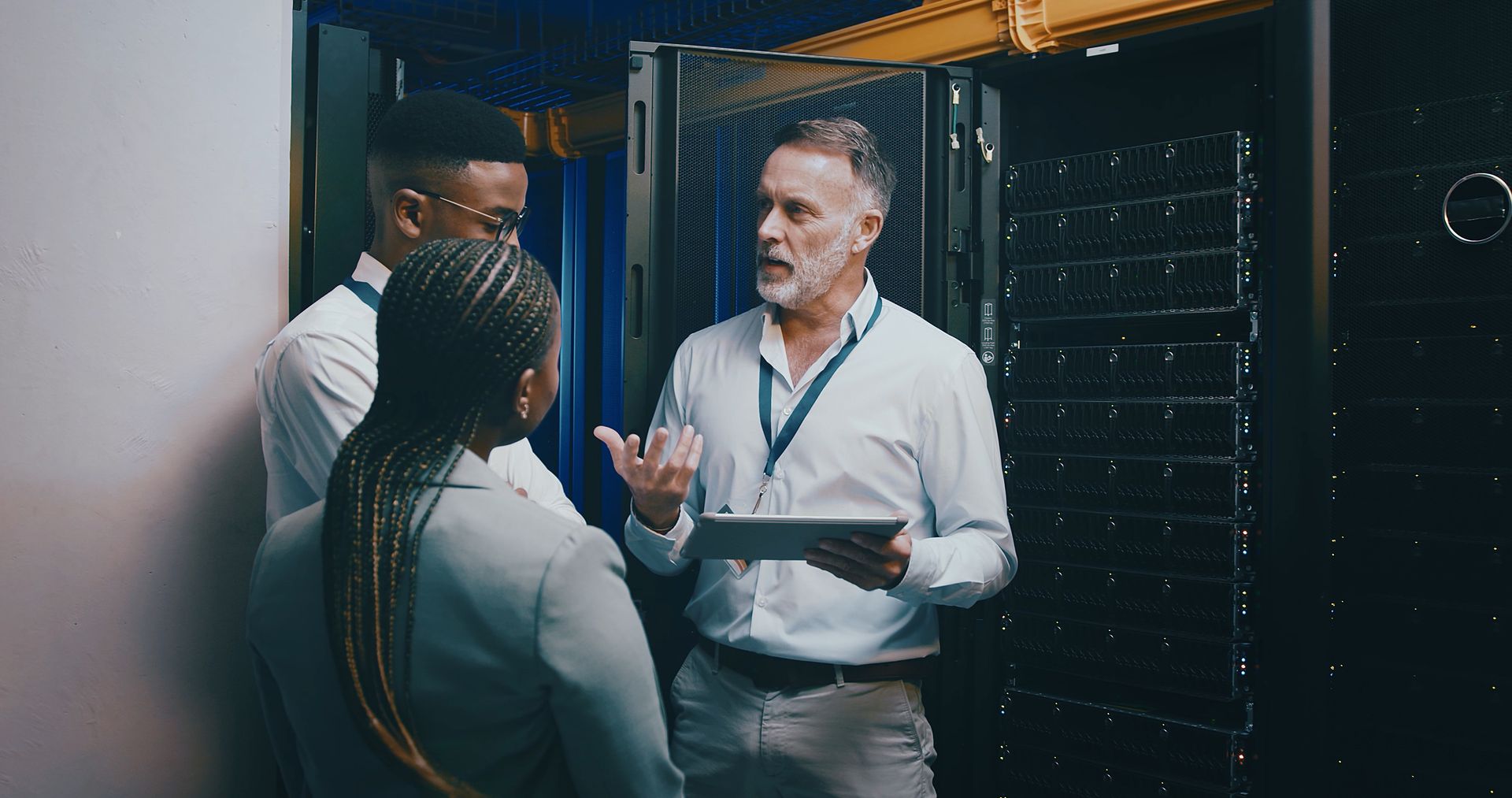 Three people discussing in a server room, with one holding a tablet and gesturing toward equipment racks.
