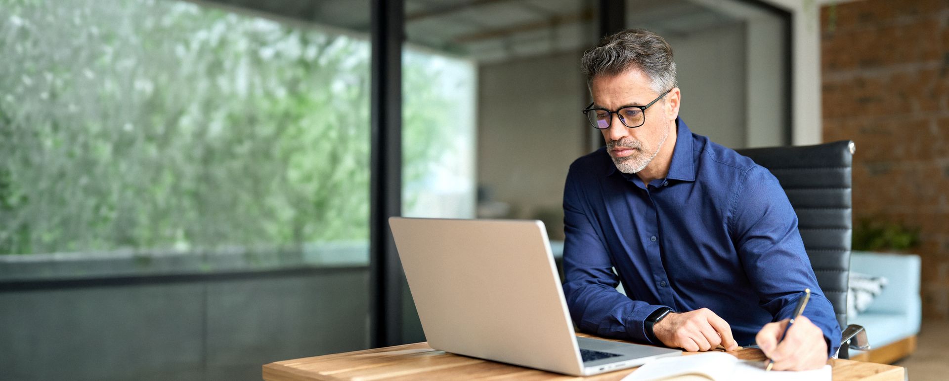 Man in a blue shirt working on a laptop at a desk by a large window