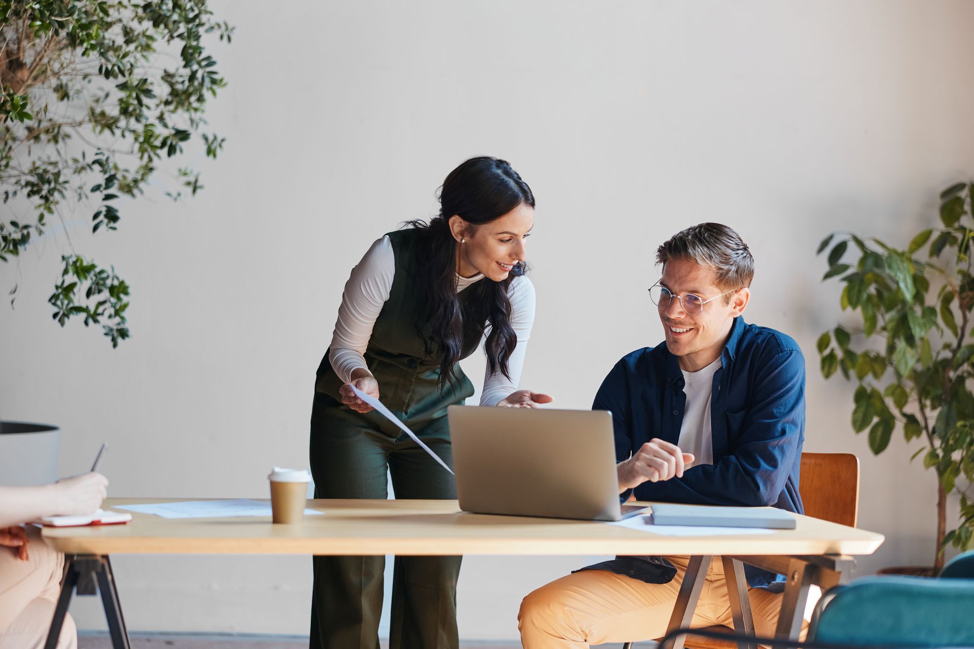 Two coworkers collaborating at a laptop in a bright office with plants and a coffee cup on the table