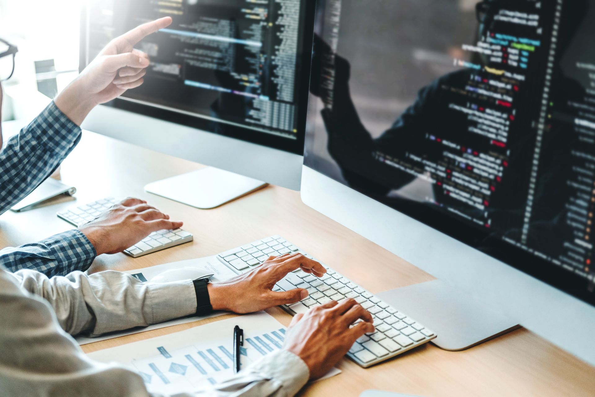 People coding at dual monitors in a bright office, hands on keyboards and screens showing code