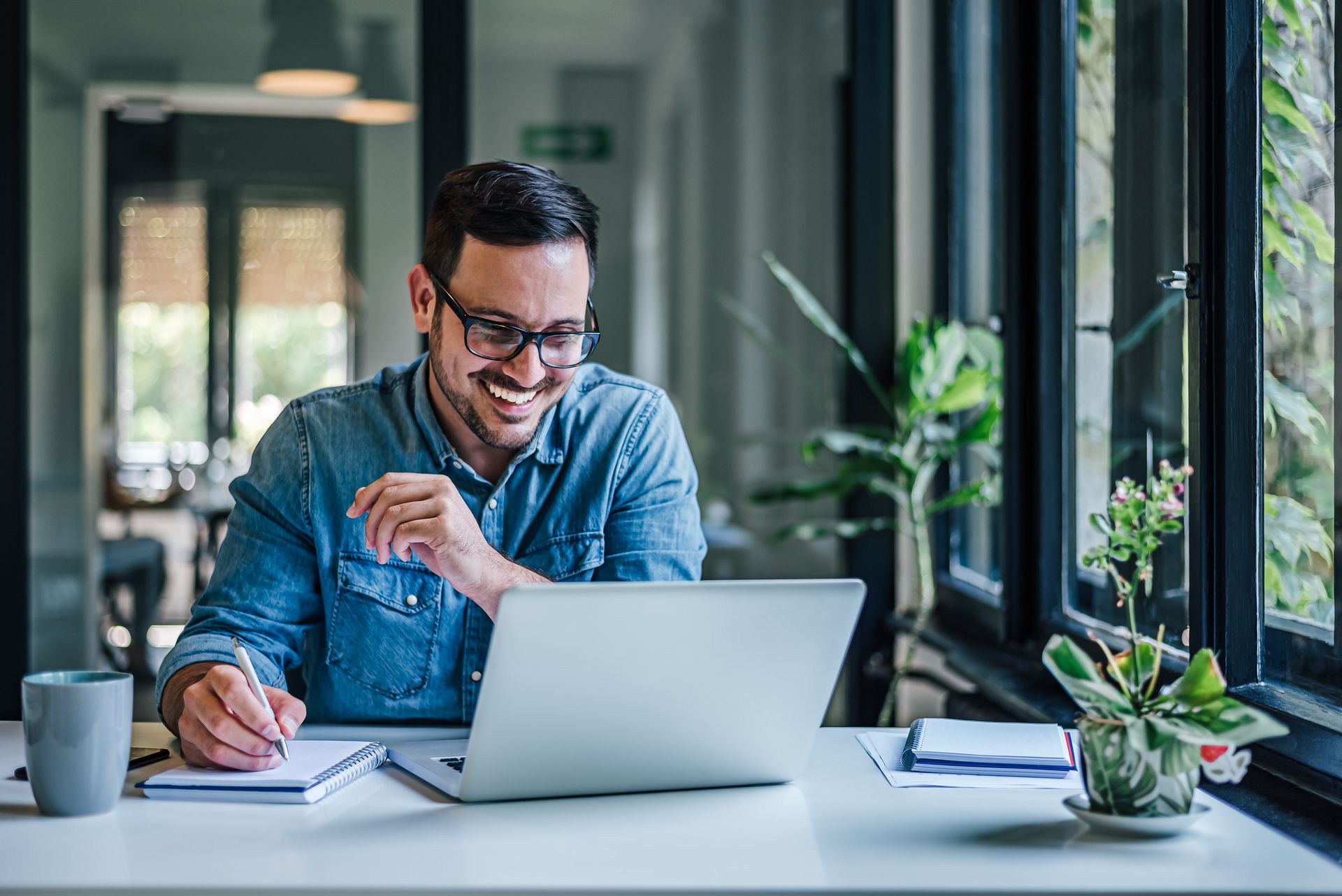 Person smiling and writing at a laptop in a bright office with plants and large windows
