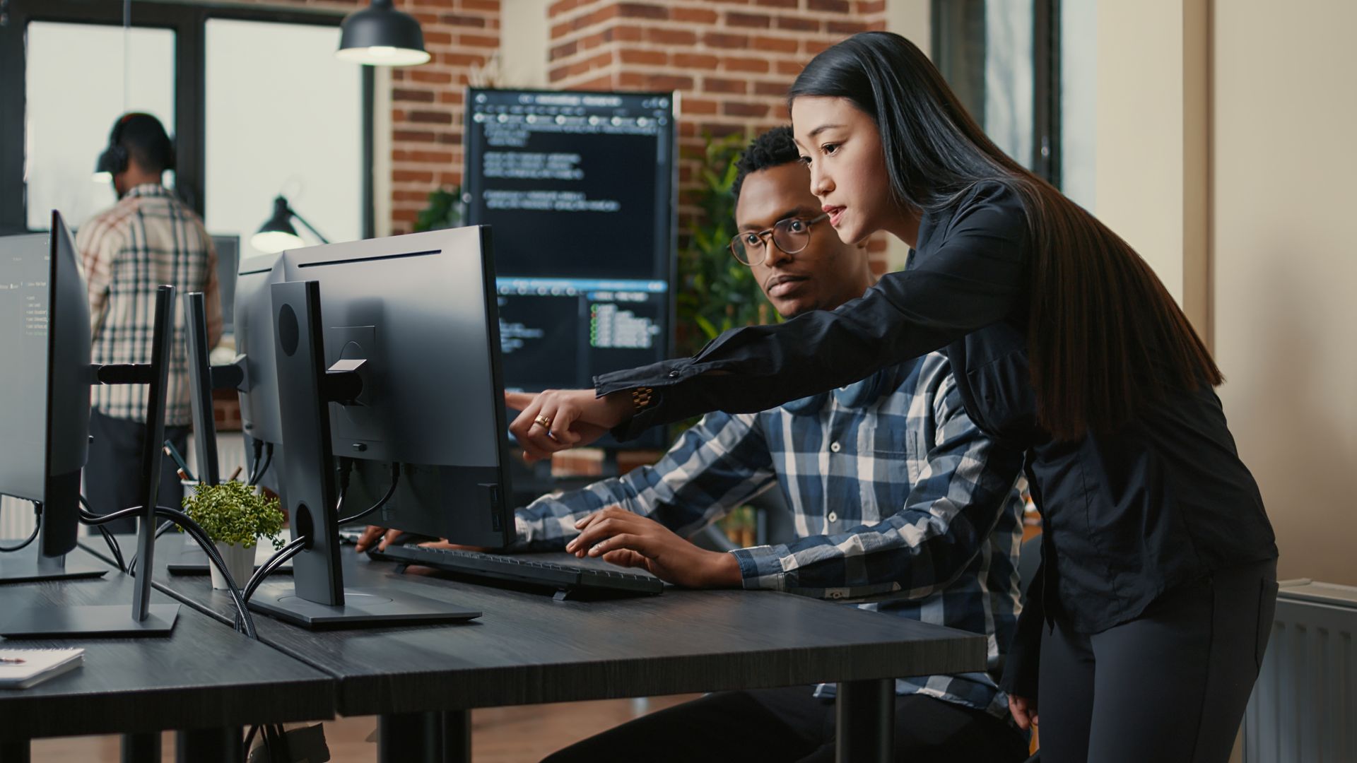 Two coworkers collaborate at a computer workstation in a modern office with monitors and a whiteboard.