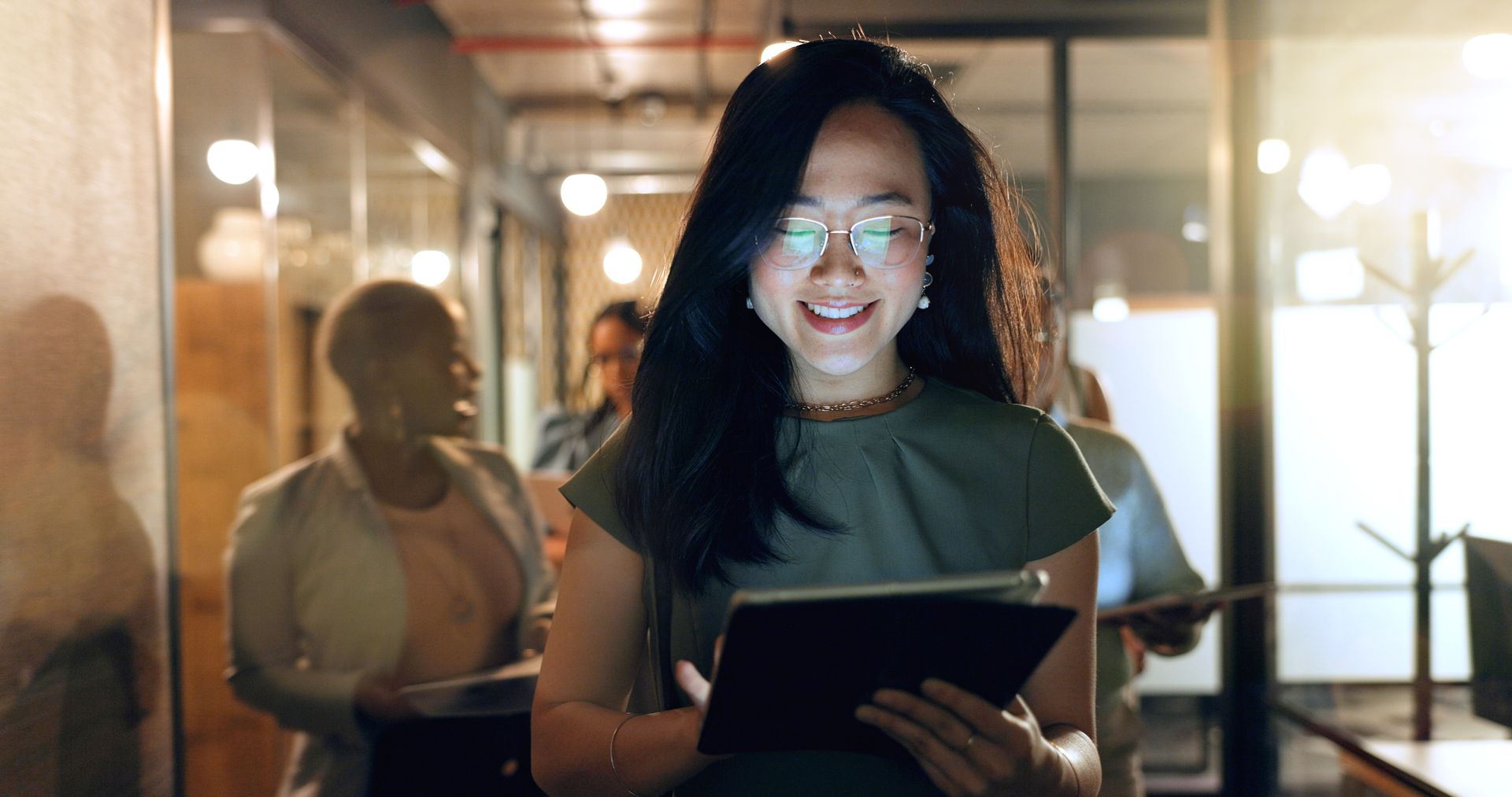 Person with glasses holding a tablet in a warmly lit office corridor, smiling at the screen