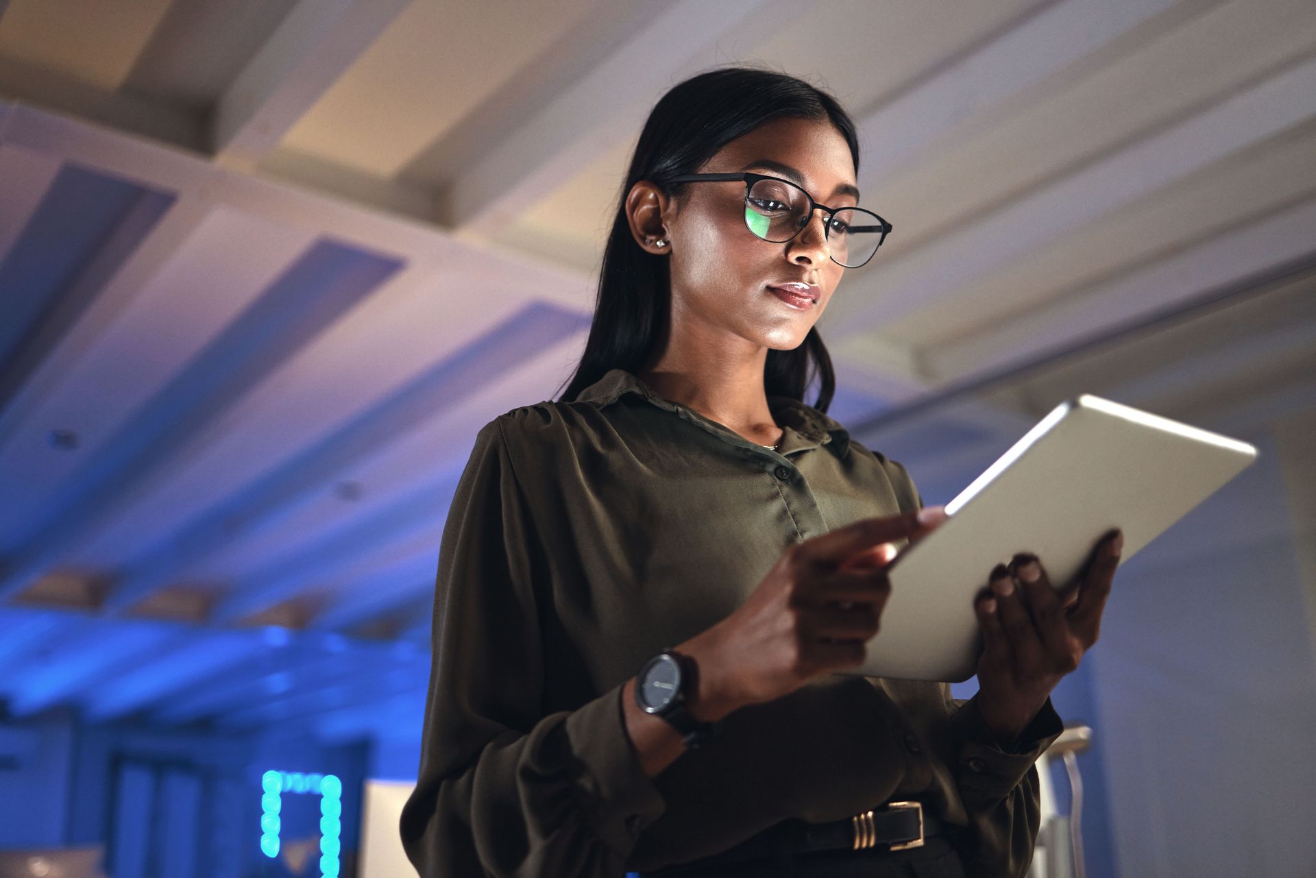 Person wearing glasses reads from a tablet in a blue-lit room.