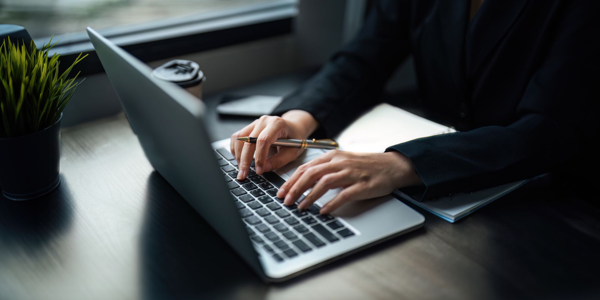 Hands typing on a laptop at a desk beside a small potted plant