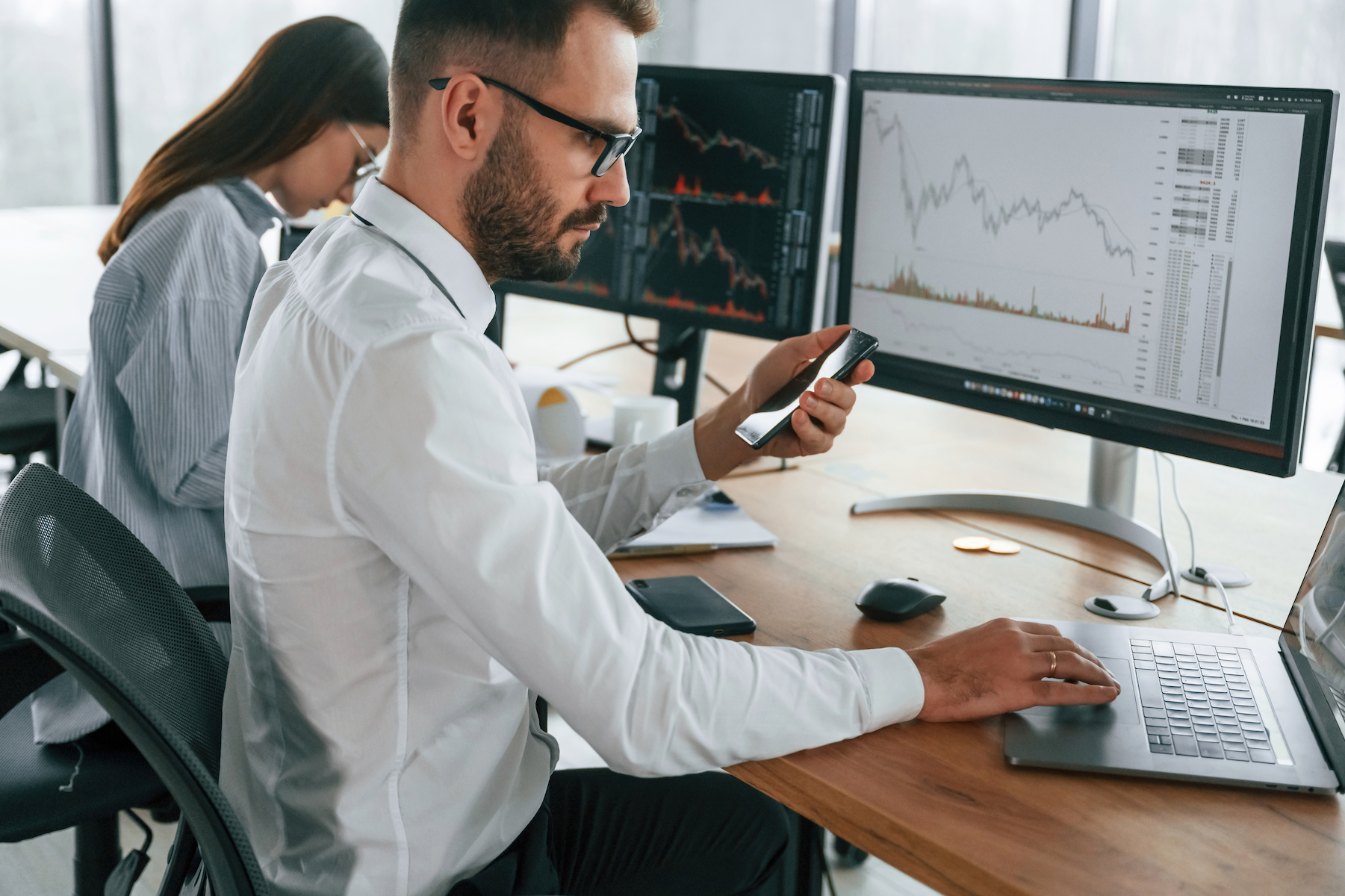 Two office workers analyzing financial charts on dual monitors, one using a phone at a desk