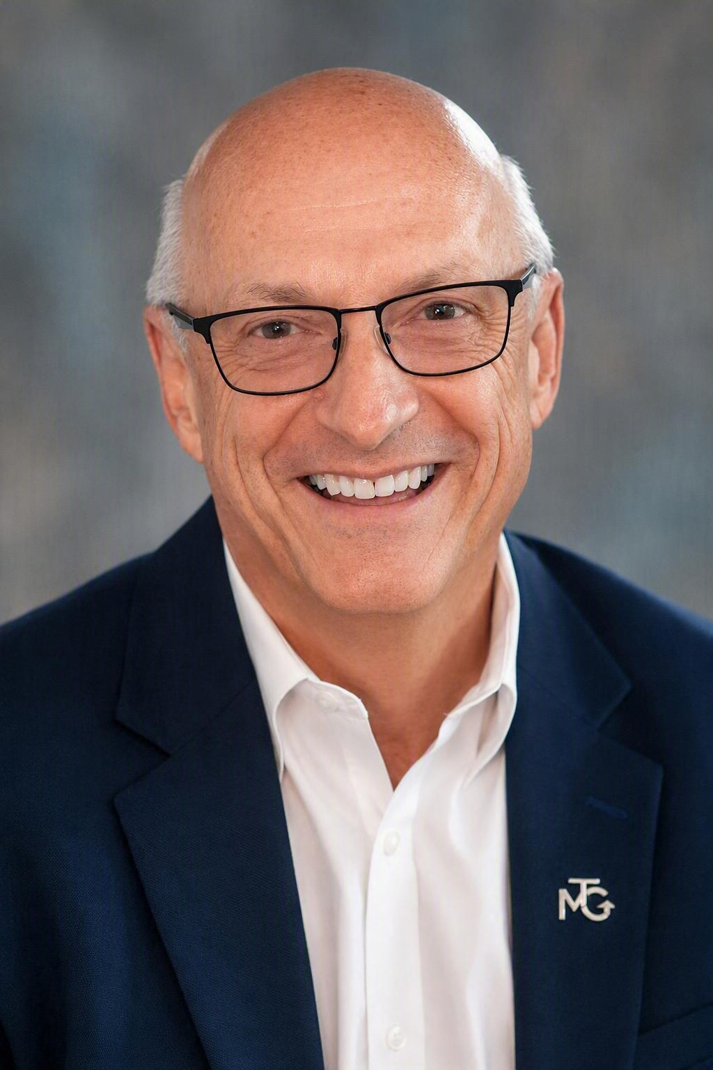 Smiling man in glasses and a navy blazer against a gray studio backdrop
