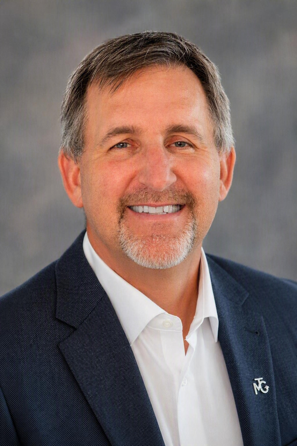 Professional headshot of a smiling man in a dark blazer and white shirt against a gray background