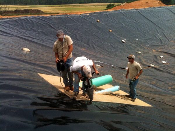 Three men working on a large black liner, possibly for a pond or landfill, using tools. One man drills a hole, while the others observe.