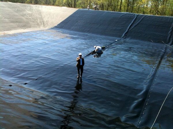 Two people in a large, dark-lined containment pond; one stands while the other works on a pipe.