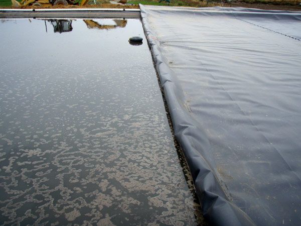 Black liner bordering water-filled containment area; a backhoe is visible in the background.