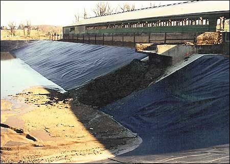 Covered bridge over a waterway with dark blue liners on the banks and a build-up of dark sediment.