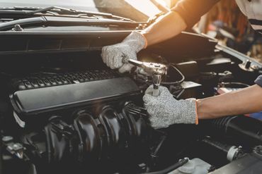 Mechanic's gloved hands working on a car engine in a garage, with the hood open.