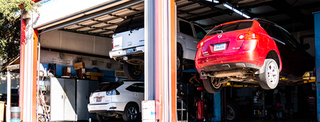 Auto repair shop with cars lifted on hydraulic lifts, including a red car and a white car inside the garage