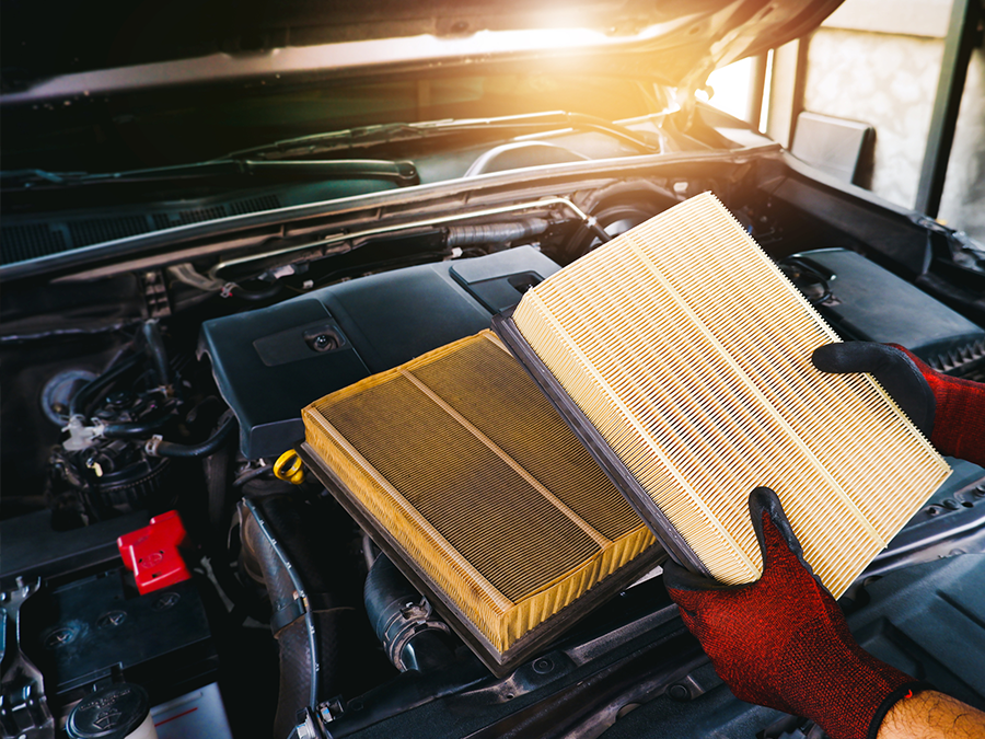 Hands holding dirty and clean car engine air filters under an open hood at sunset