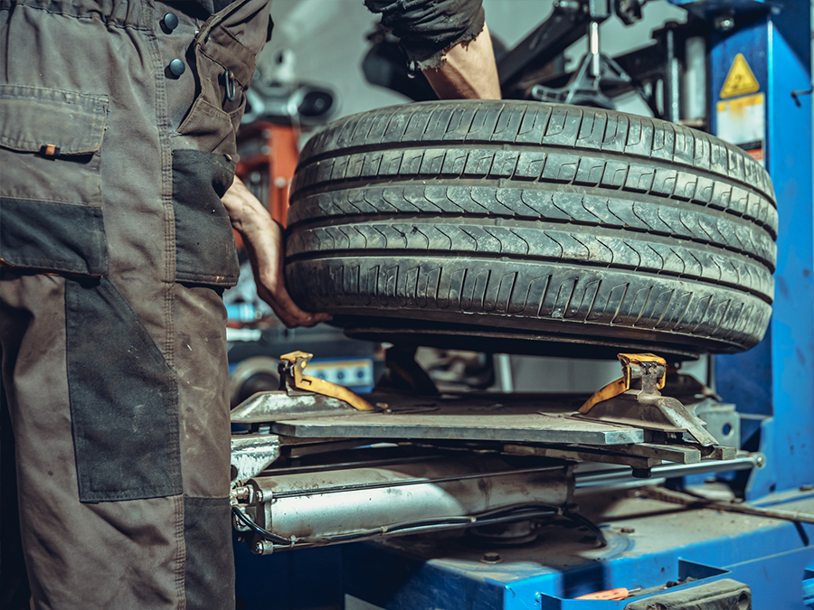 Mechanic working under a car on a lift, with stacked tires visible in the garage.