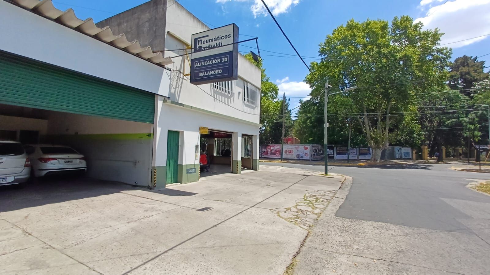 Esquina soleada con un edificio blanco, aparcamiento cubierto, letrero y árboles a lo largo de una calle tranquila.