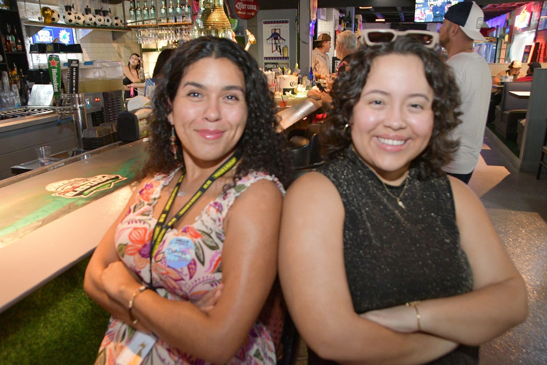 Two women are standing back to back in a bar with their arms crossed.