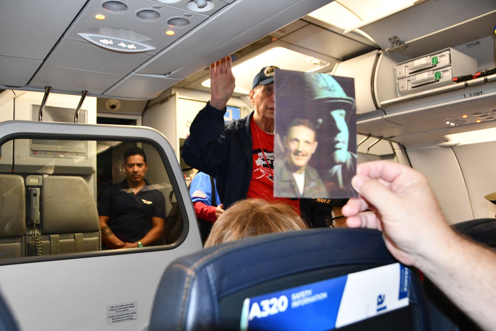 Man on plane raises hand, holding old photo. Flight attendant watches.