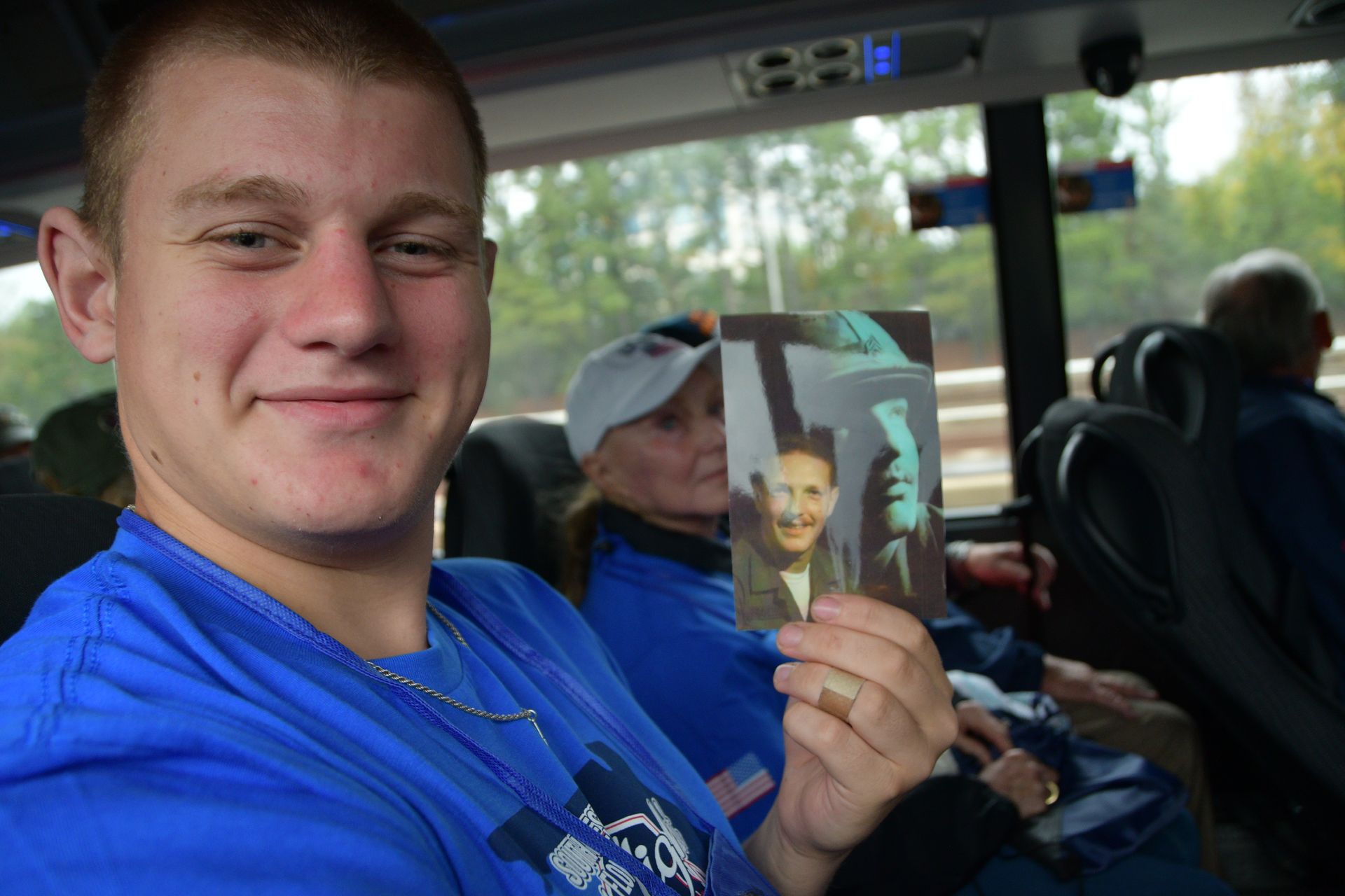 Man on a bus smiles, holding a photo of a child. Passengers and windows visible.