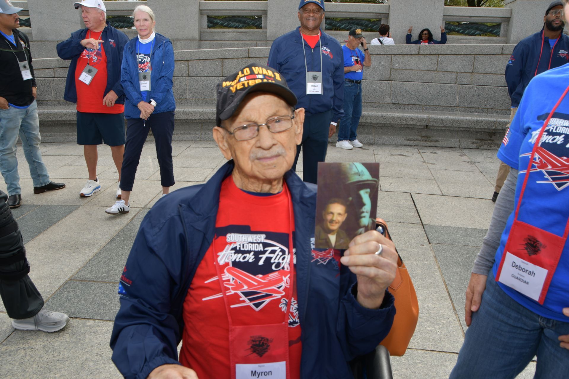 Elderly man in a wheelchair holds a photo. He wears a red shirt, blue jacket, cap, and glasses. Group in background.