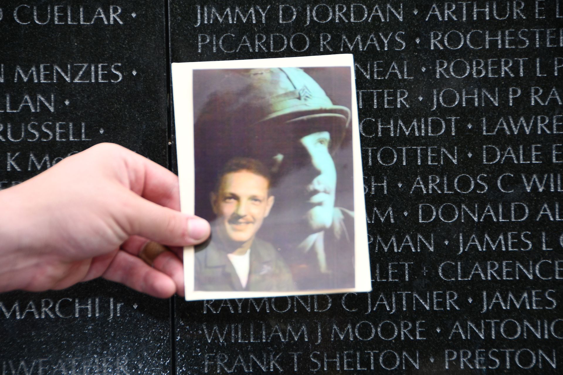 Hand holding a photo of a man at the Vietnam Veterans Memorial, names etched in black granite.