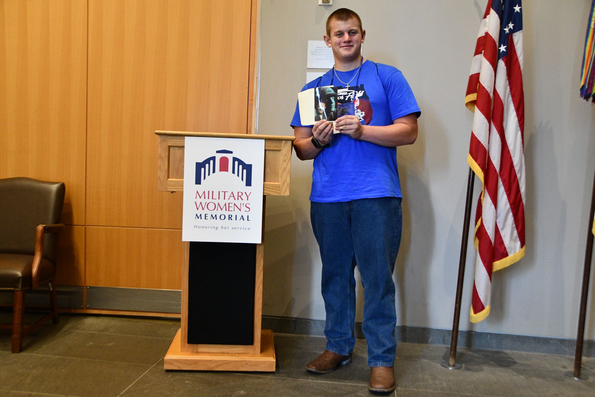 Man in blue shirt and jeans holds book at a podium, next to American flag.