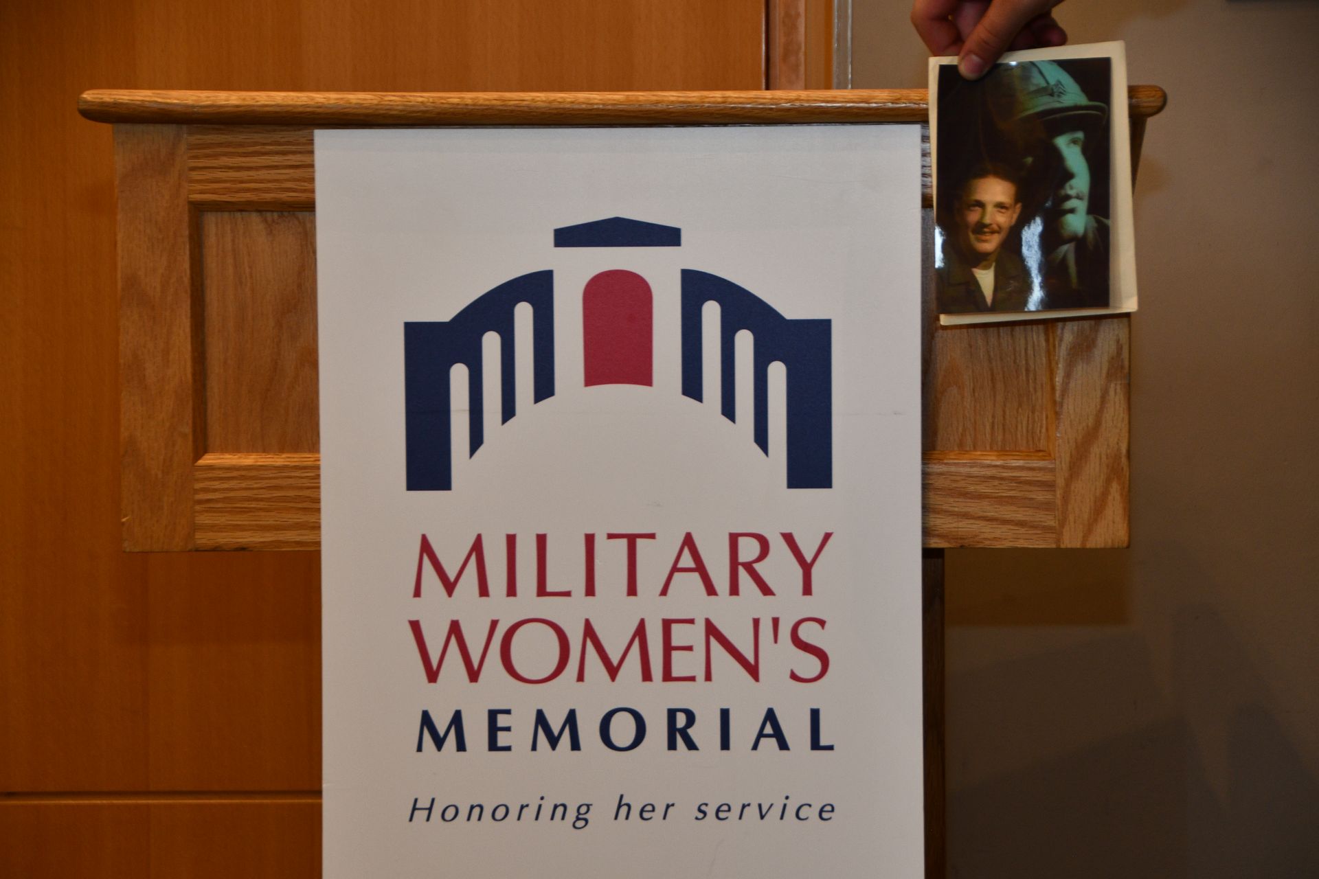 Military Women's Memorial sign; a person holds up a photo. The sign is white with blue and red text and an arch design.