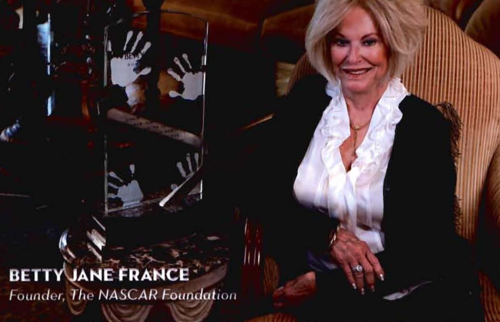 Betty Jane France seated indoors, smiling in a white blouse and dark jacket, with text identifying her as NASCAR Foundation founder