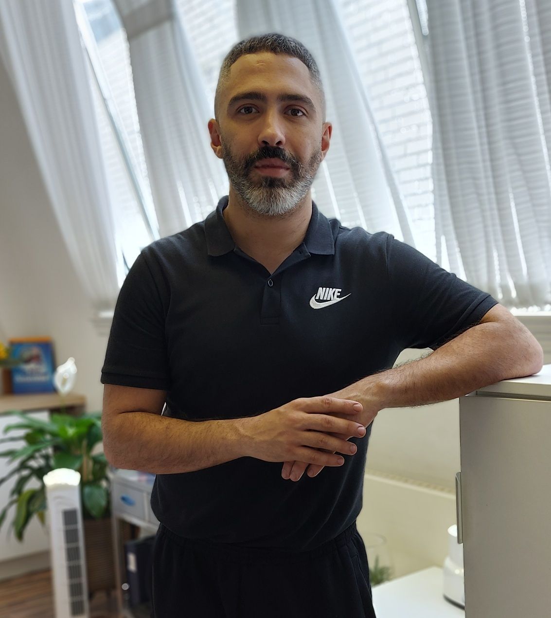 Manil, a Sports Therapist at R P Sports Clinic, is standing confidently in the clinic wearing a black Nike polo shirt.