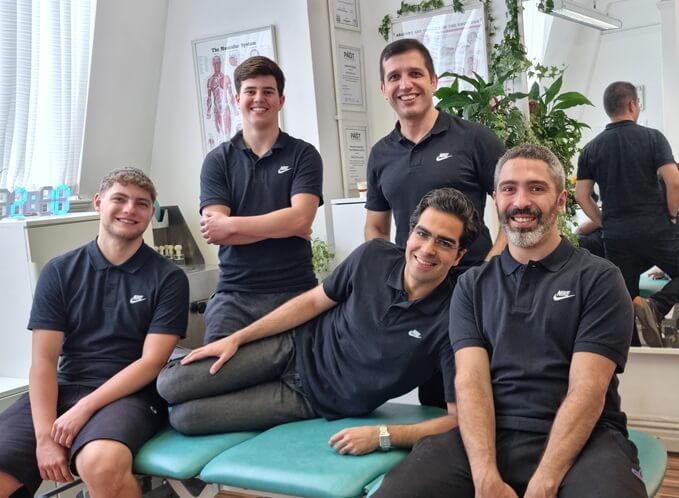 A group of five expert sports therapists at R P Sports Injuries & Wellness Clinic smiling together in a treatment room.