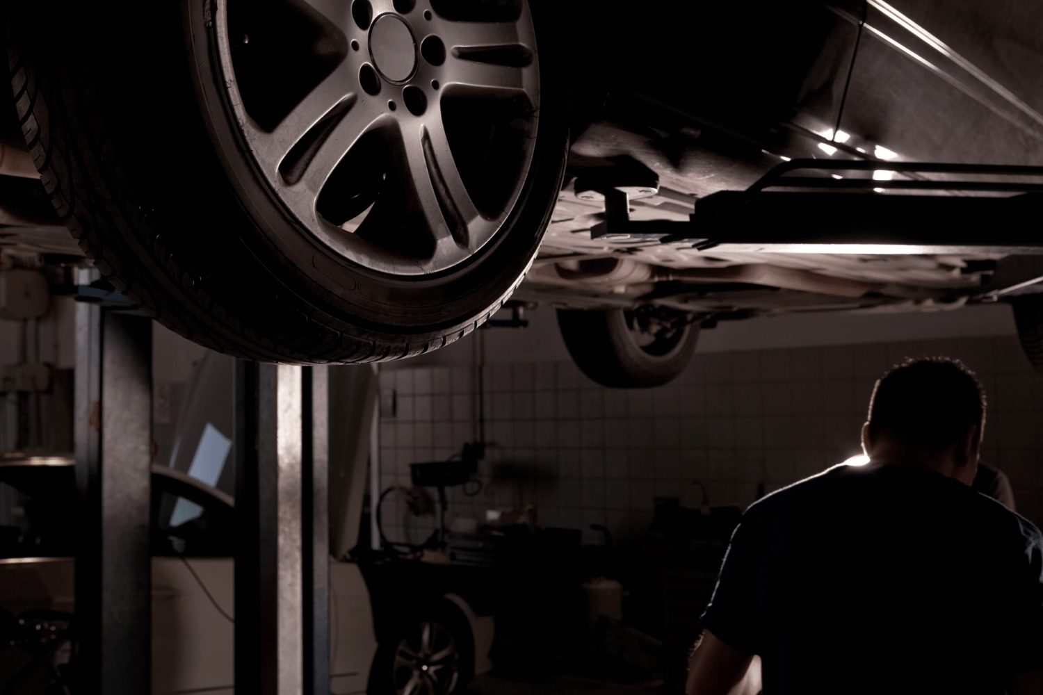 Car lifted in an auto repair shop, with a mechanic working in the dim background.