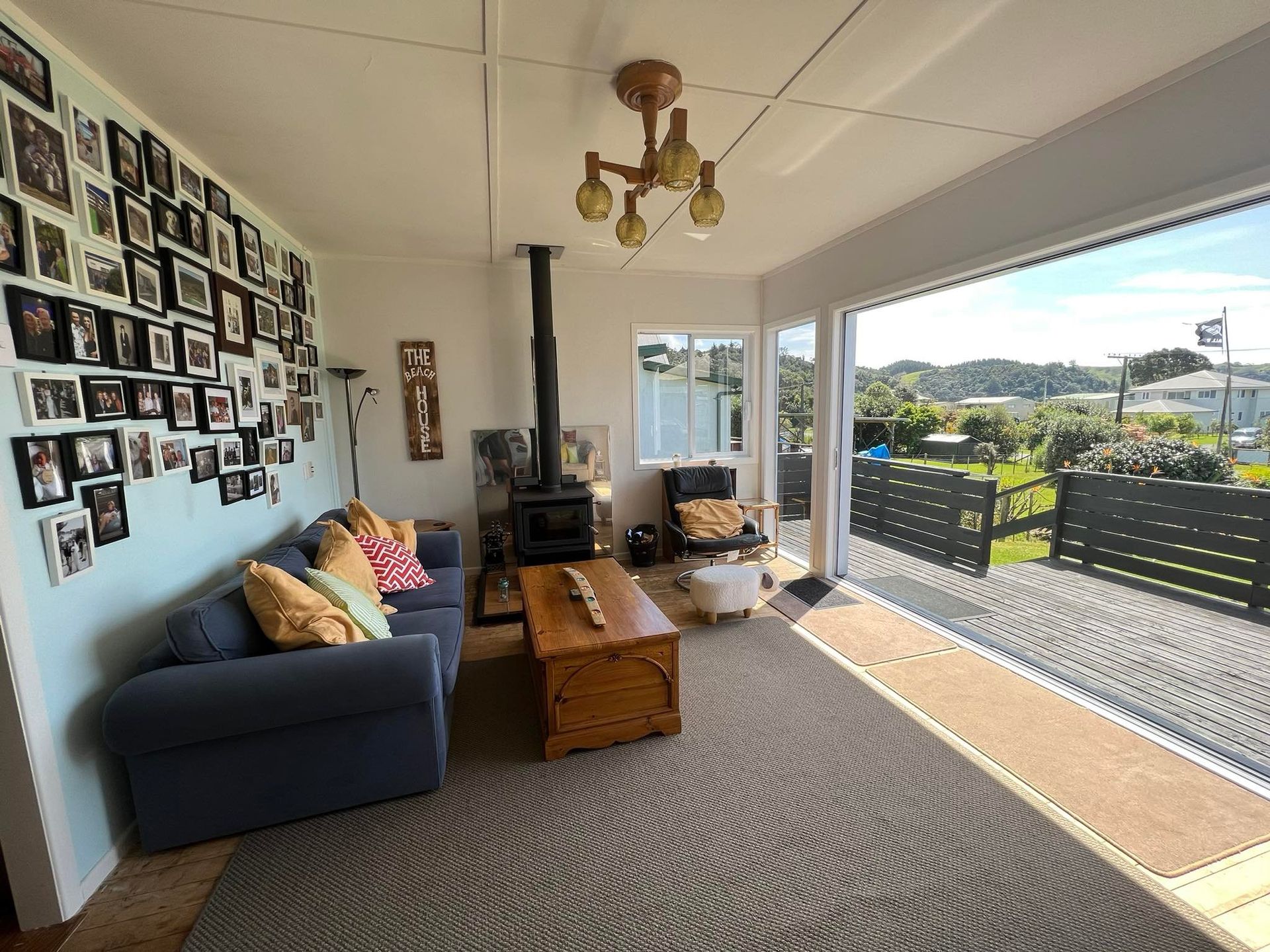 Cozy living room with gray sofa, wood stove, photo wall, and sliding doors opening to a bright deck and hillside view