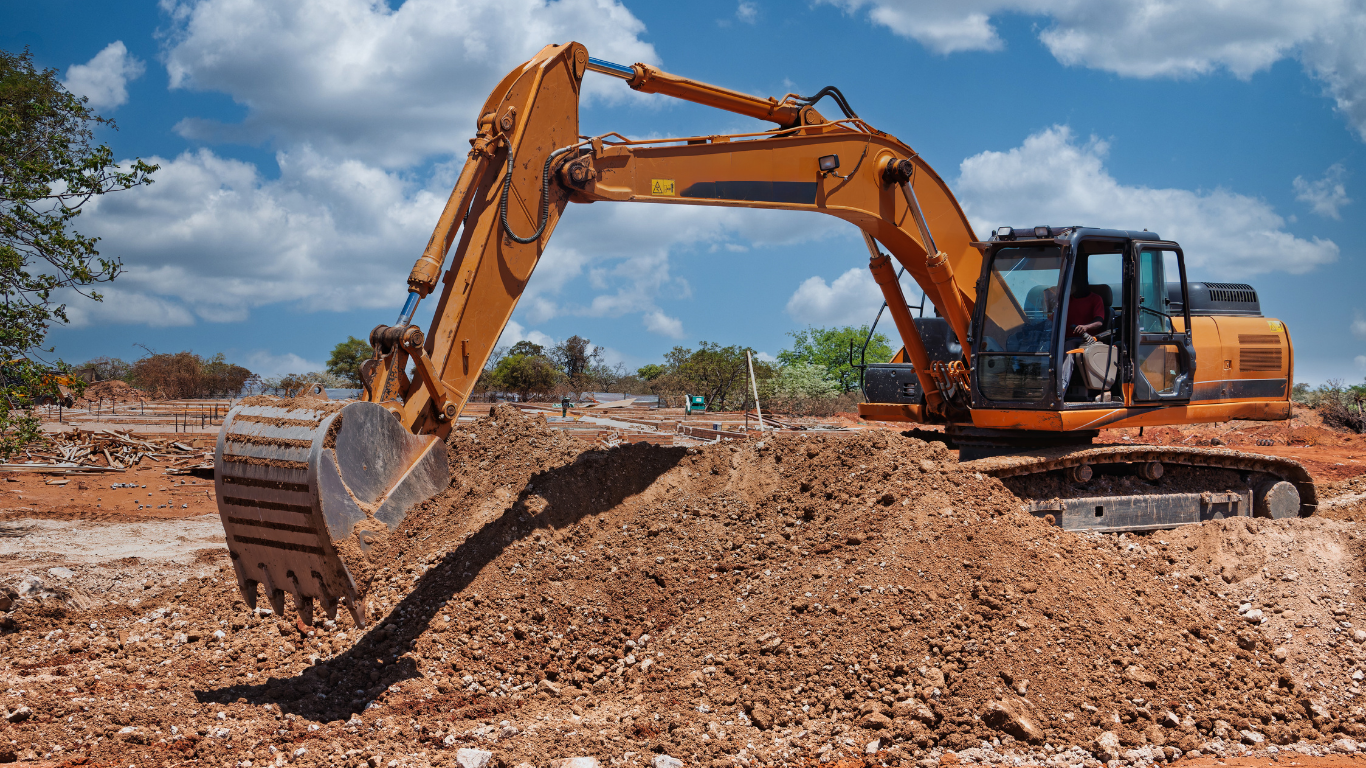 An orange excavator scoops dirt on a construction site under a blue sky with white clouds.