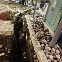 A construction trench runs alongside a stone-filled border, with a utility box and post visible in the background.