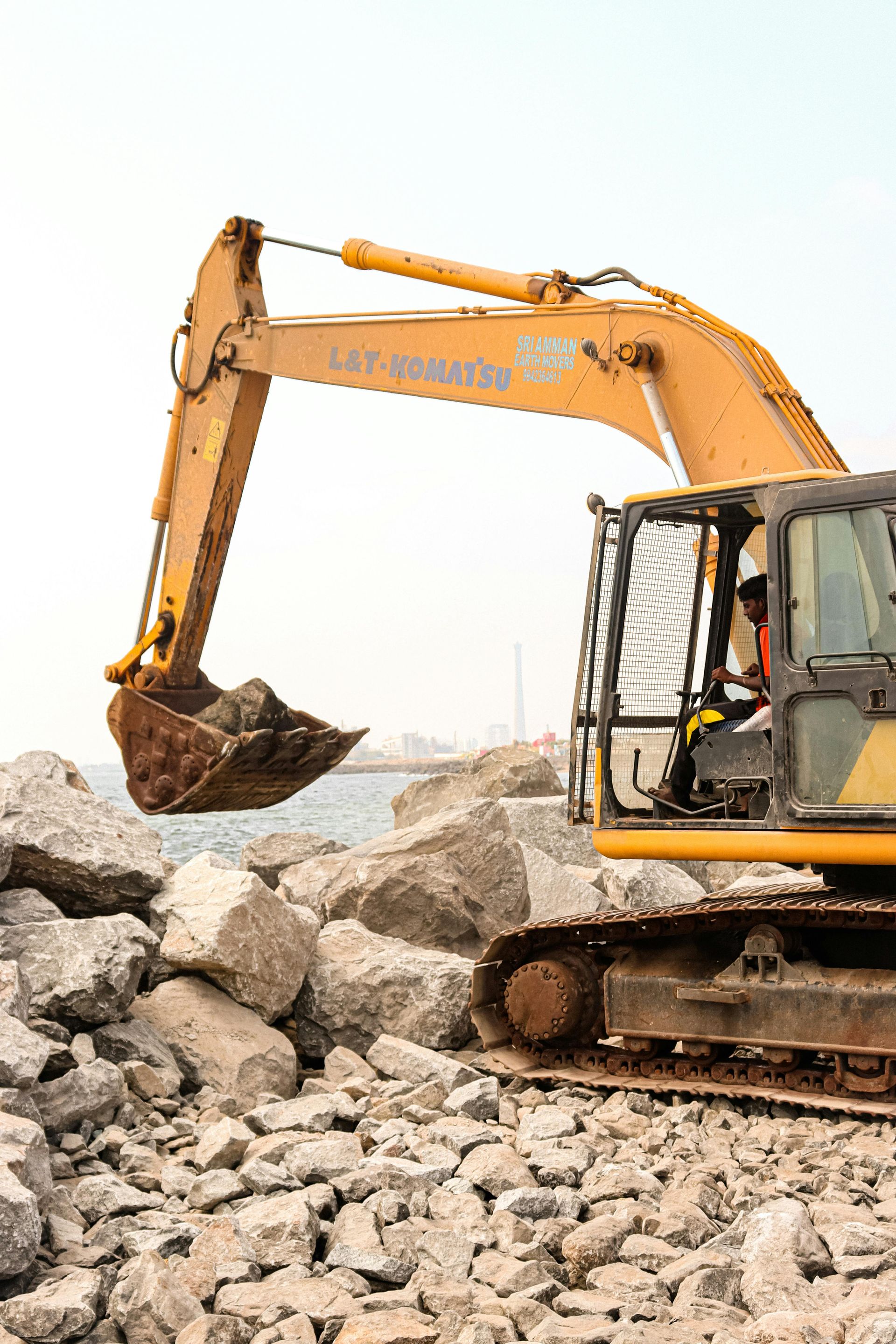 A yellow excavator lifts a large rock on a construction site near a body of water.