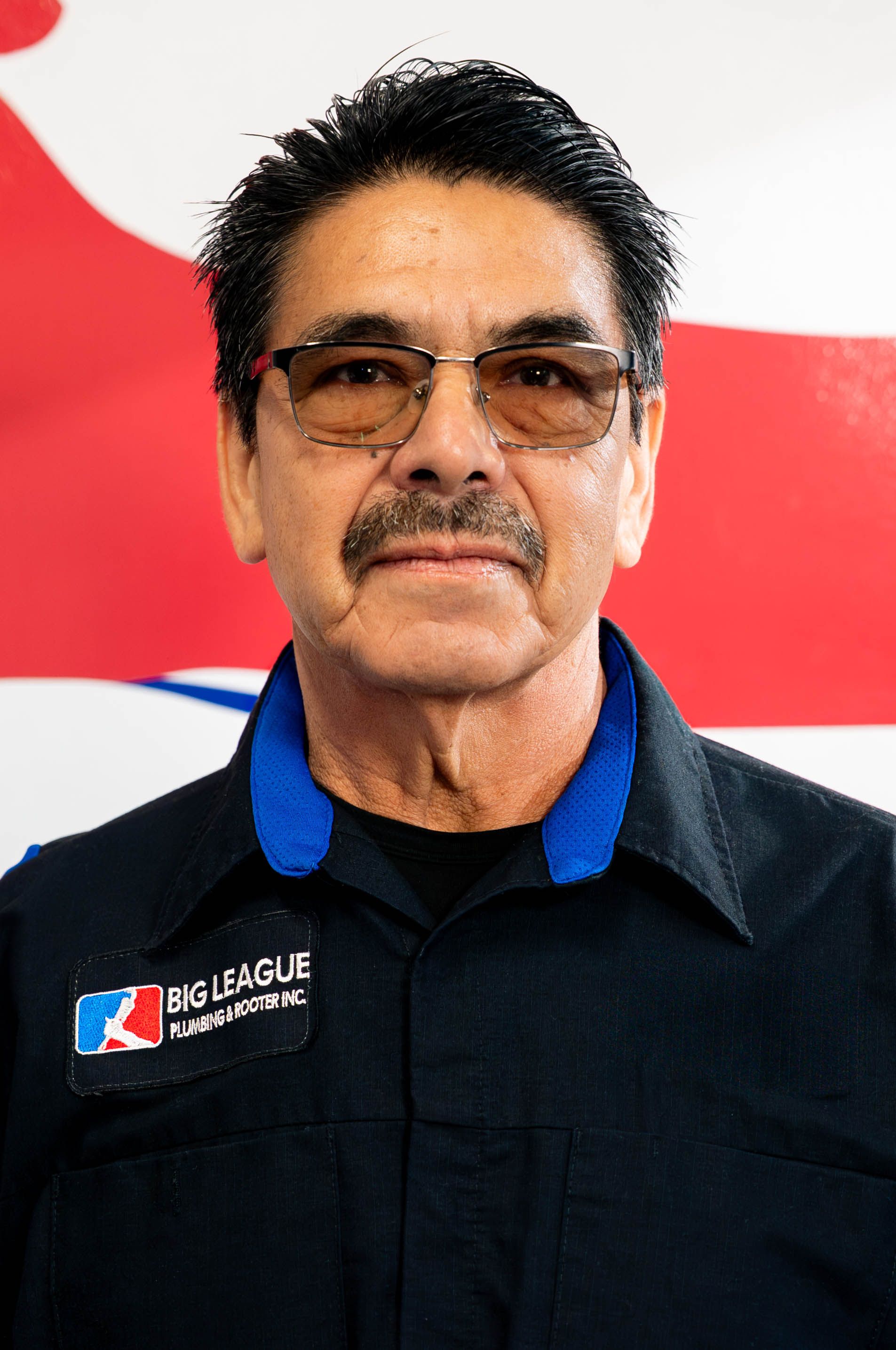 Man wearing sunglasses and black shirt with a logo, standing before a red-and-white backdrop