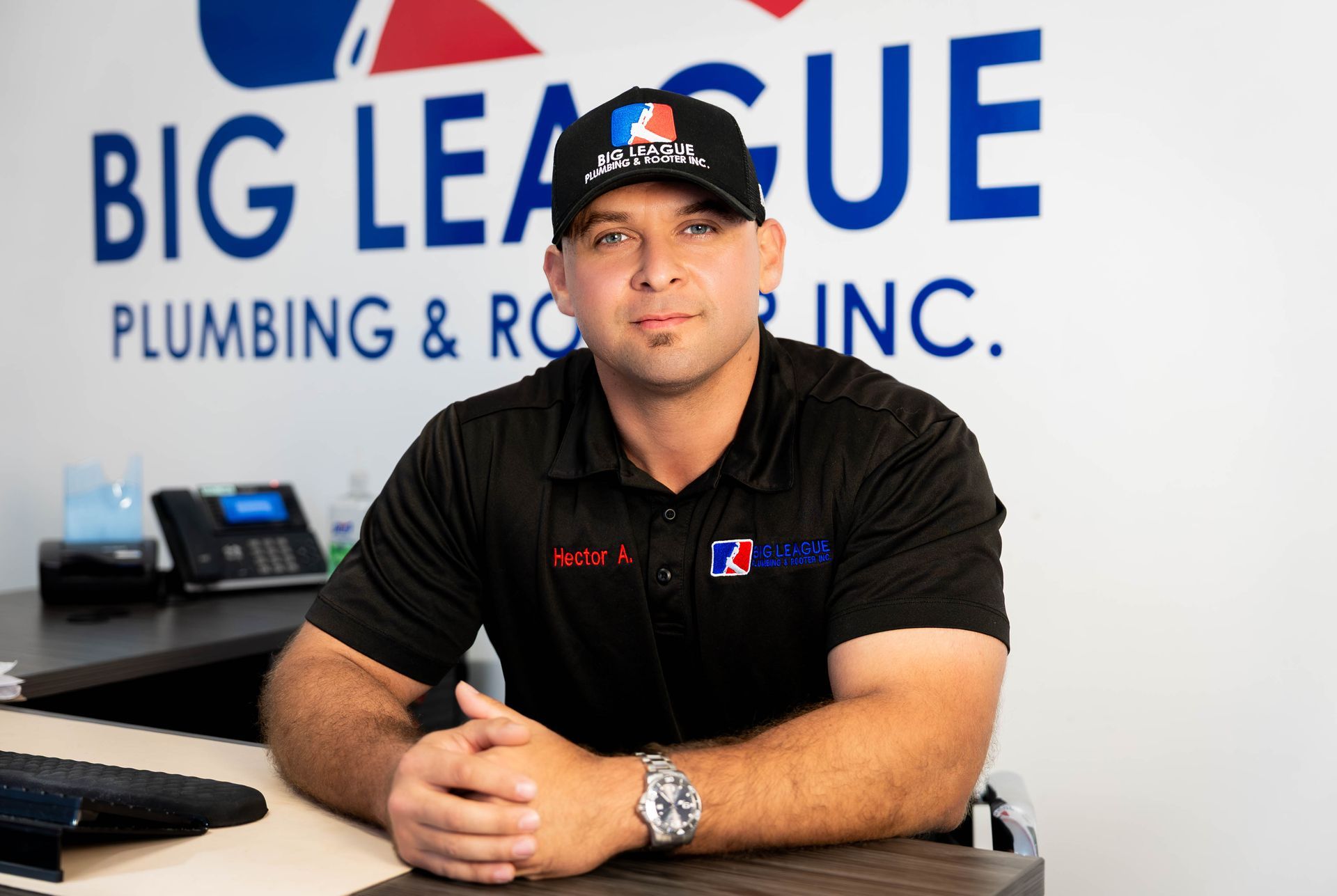 Man seated at a desk in front of Big League Plumbing & Rooter Inc. sign, wearing a black cap and shirt