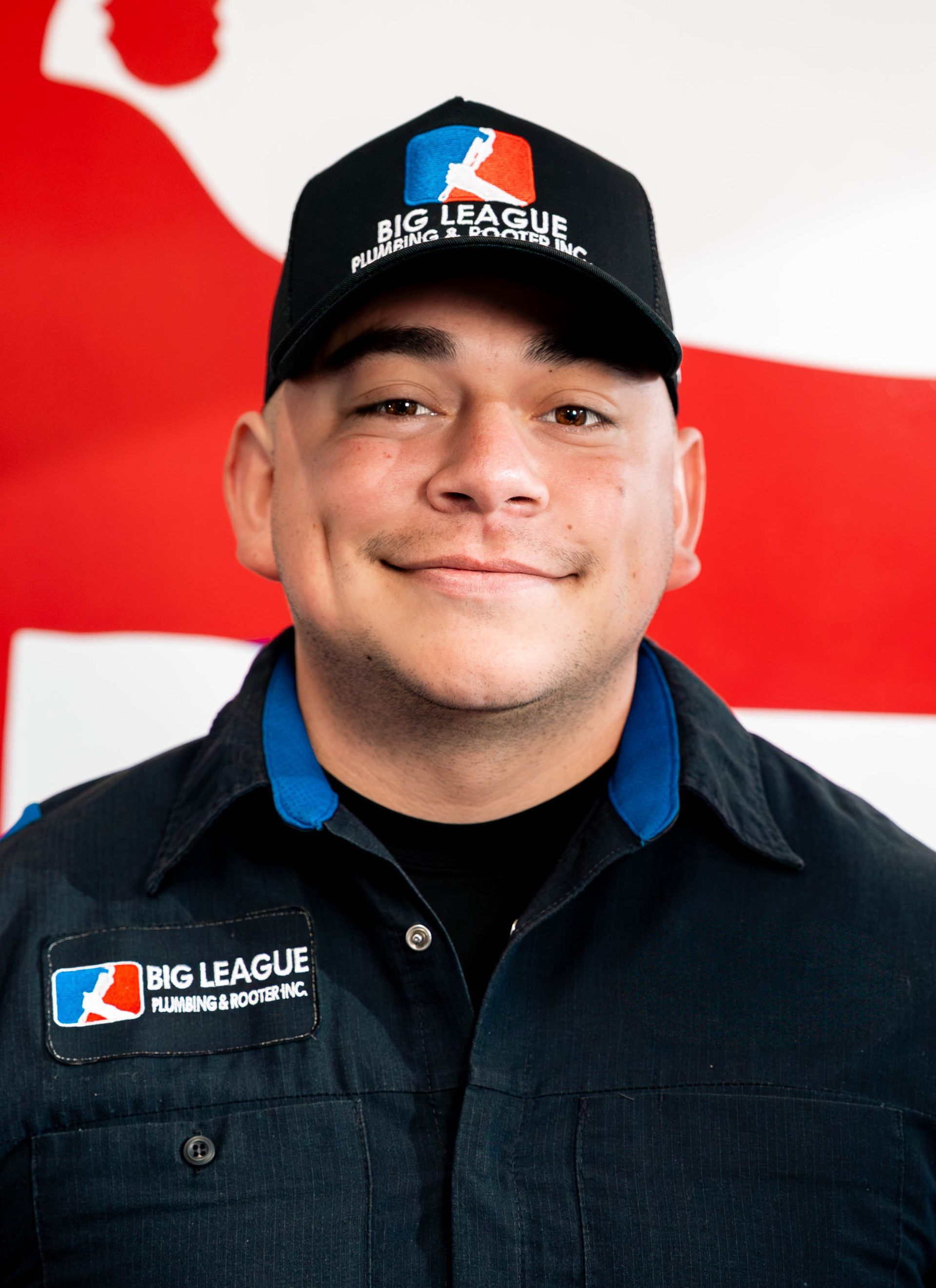 Smiling MLB League employee in navy jacket and black cap, standing before a red-and-white flag backdrop