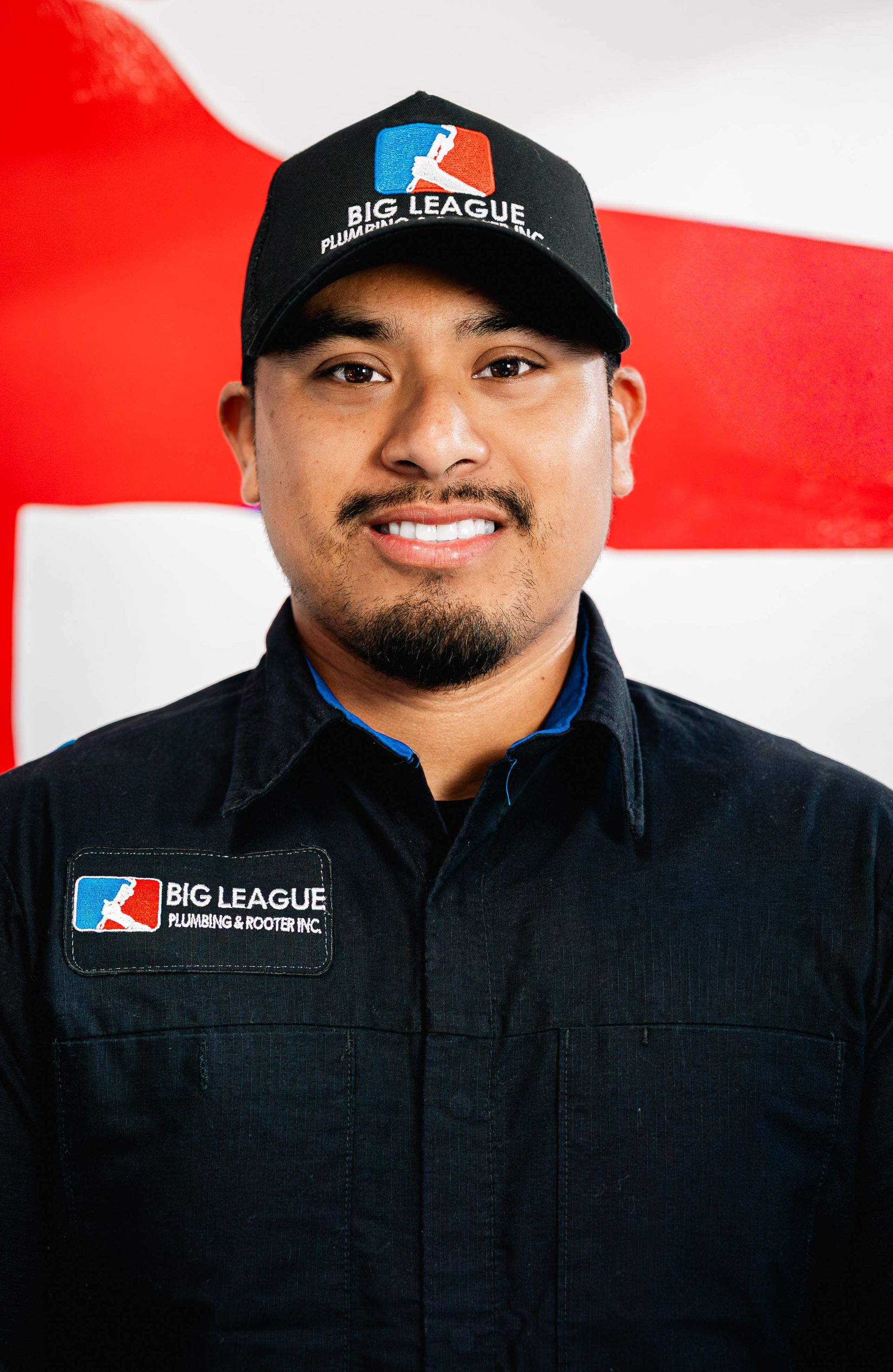 Portrait of a smiling person in a black cap and shirt with a Big League logo, against a red and white backdrop