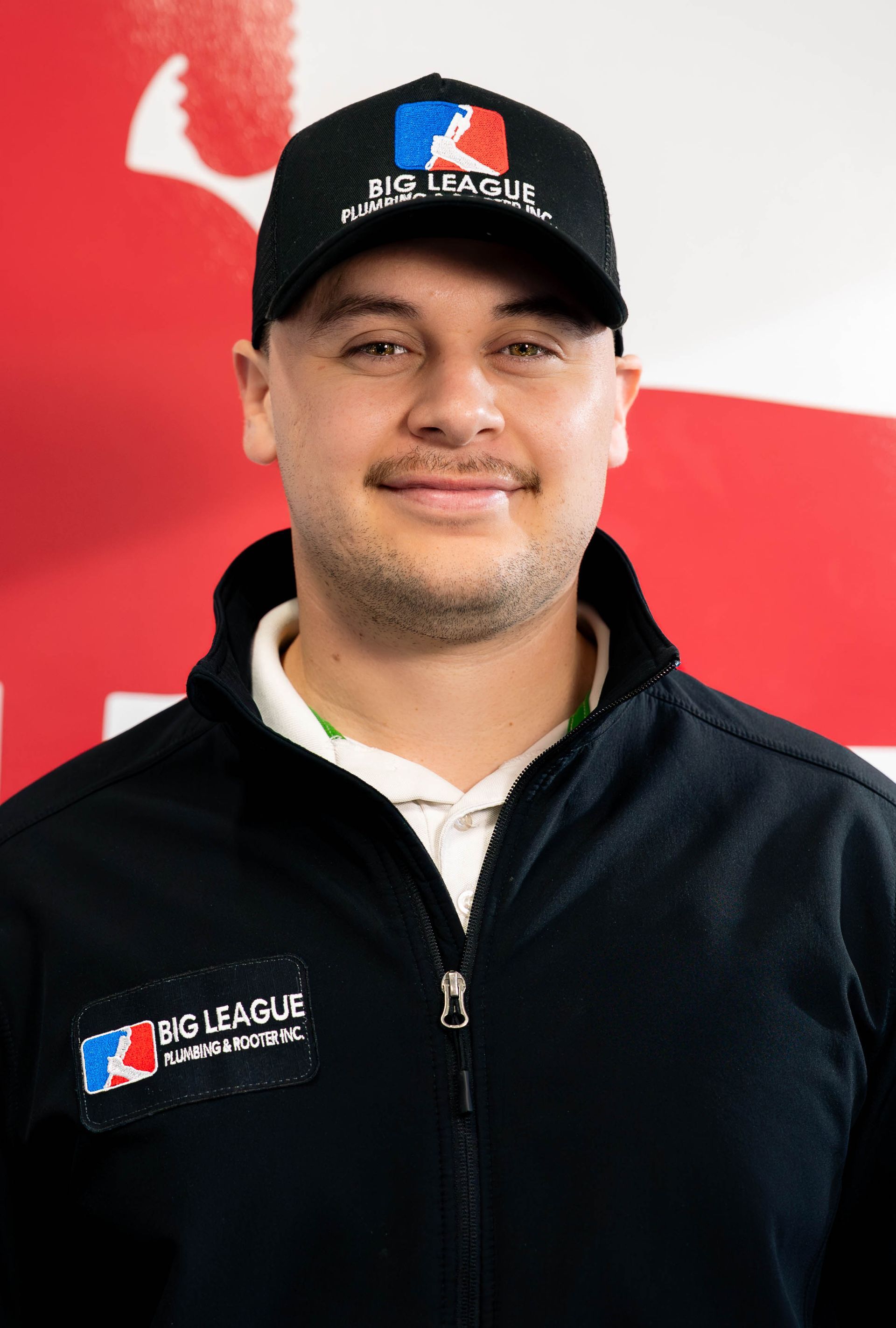 Man in a black cap and jacket with a logo, standing against a red-and-white backdrop.